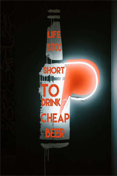 Φωτιζόμενο μπύρα μπουκάλι σύμβολο στο σκοτάδι τείχος στην παμπ με την επιγραφή «η ζωή είναι πολύ μικρή για να πιείτε φτηνή μπύρα» - Φωτογραφία, εικόνα