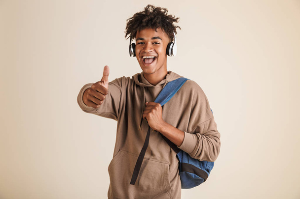 Portrait of a cheerful young afro american man dressed in hoodie with headphones carrying backpack isolated, showing thumbs up - Valokuva, kuva