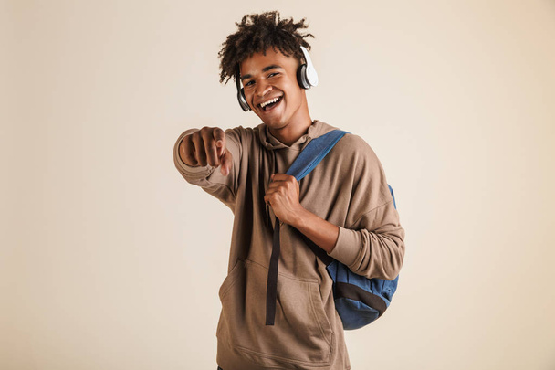 Portrait of a cheerful young afro american man dressed in hoodie with headphones carrying backpack isolated, pointing at camera - Foto, Bild