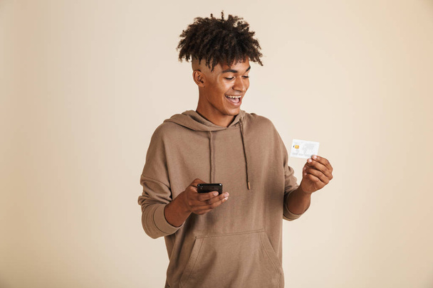 Portrét usměvavé mladé afro americký muž oblečený v hoodie izolovaná, pomocí mobilního telefonu, ukazující plastové platební karty - Fotografie, Obrázek