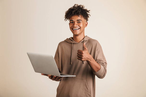 Portrait of a smiling young afro american man dressed in hoodie isolated, using laptop computer, showing thumbs up - Photo, Image