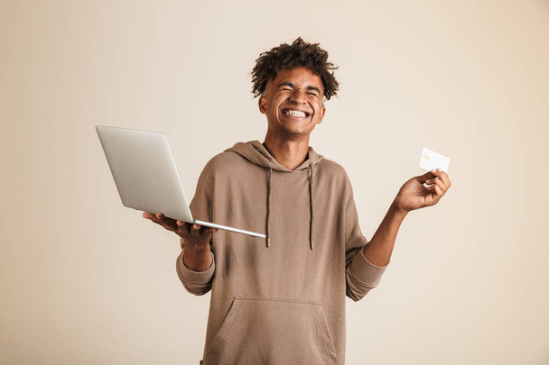 Portrait of a joyful young afro american man dressed in hoodie isolated, using laptop computer, showing plastic credit card - Foto, Imagen
