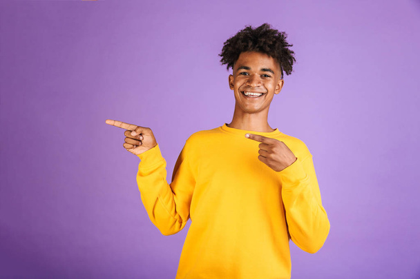 Portrait of a smiling young afro american man dressed in sweatshirt pointing fingers away at copy space isolated - Foto, Bild
