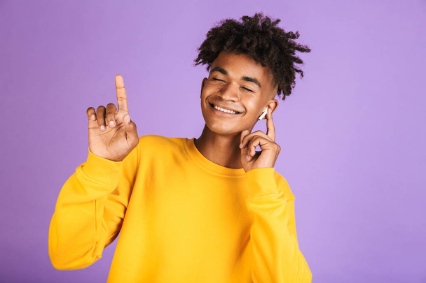 Portrait of happy african man having stylish afro hairdo dancing while listening to music via bluetooth earphone, isolated over violet background - Photo, image