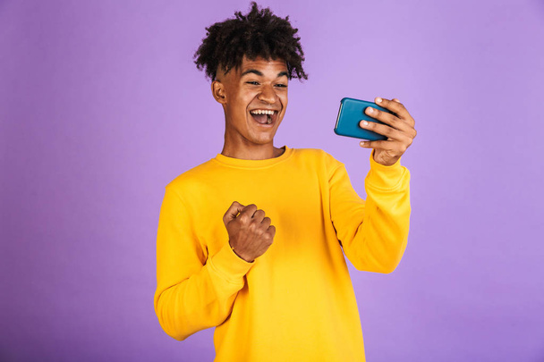 Portrait of a satisfied young afro american man dressed in hoodie isolated, playing games on mobile phone - Foto, afbeelding