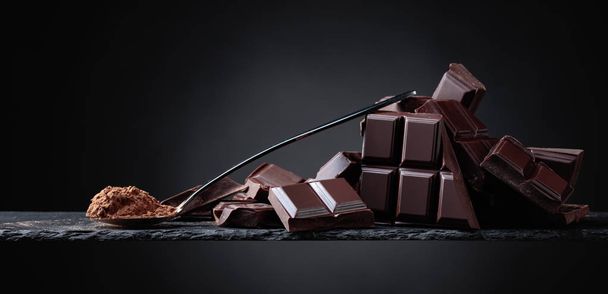 Broken chocolate pieces and cocoa powder in small spoon on a black background. Copy space . - Photo, Image