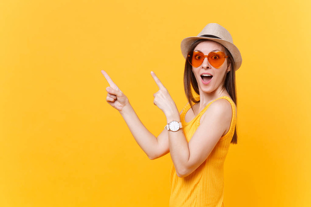 Portrait de jeune femme souriante en chapeau d'été en paille, lunettes orange pointant index de côté espace de copie isolé sur fond jaune. Les gens émotions sincères, concept de style de vie. Espace publicitaire
 - Photo, image