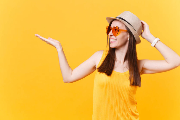 Portrait de jeune femme souriante en chapeau d'été en paille, lunettes orange pointant à droite espace de copie isolé sur fond jaune. Les gens émotions sincères, concept de style de vie. Espace publicitaire
 - Photo, image