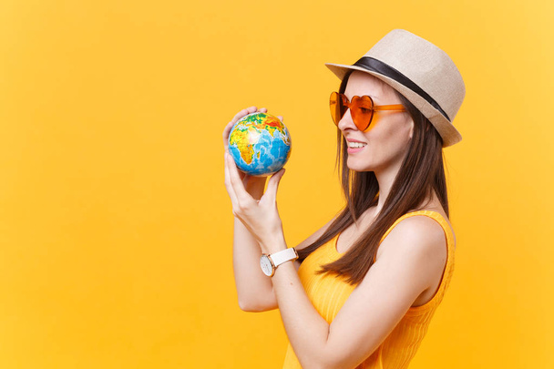 Portrait of tourist woman in summer hat, orange glasses hold in hands globe isolated on yellow background. Traveling abroad to travel weekends getaway. Air flight journey concept. Stop nature garbage - Photo, Image