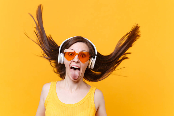 Portrait of excited cheerful laughter funny comic woman in orange glasses in headphones with fluttering hair isolated on yellow background. People sincere emotions lifestyle concept. Advertising area - 写真・画像