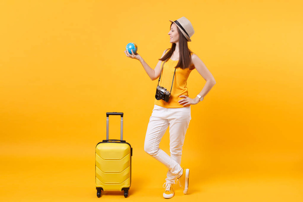 Reisende Touristin in sommerlicher Freizeitkleidung, Hut mit Globus, Koffer isoliert auf gelb-orangefarbenem Hintergrund. Passagiere, die ins Ausland reisen, um am Wochenende zu reisen. Flugreise-Konzept - Foto, Bild