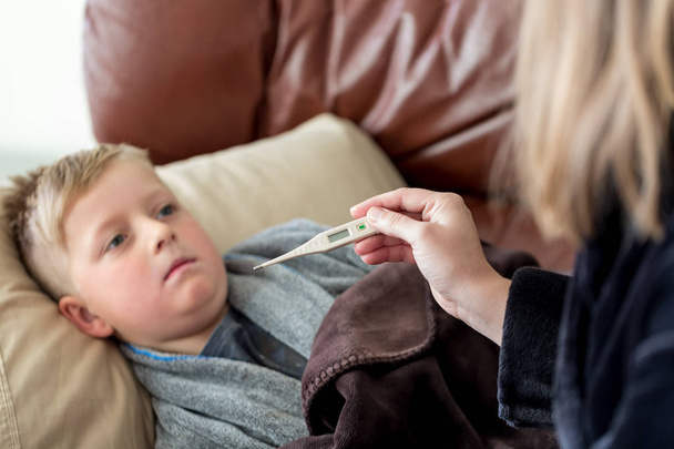 young boy laying on the couch sick while mother checks him for a fever with a thermometer - Foto, immagini