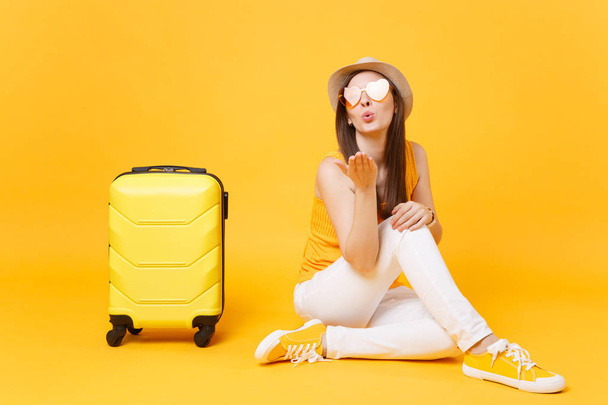 Viajero mujer turista en ropa casual de verano, sombrero sentarse con maleta aislada sobre fondo amarillo anaranjado. Pasajera viajando al extranjero para viajar los fines de semana. Concepto de viaje aéreo
 - Foto, Imagen