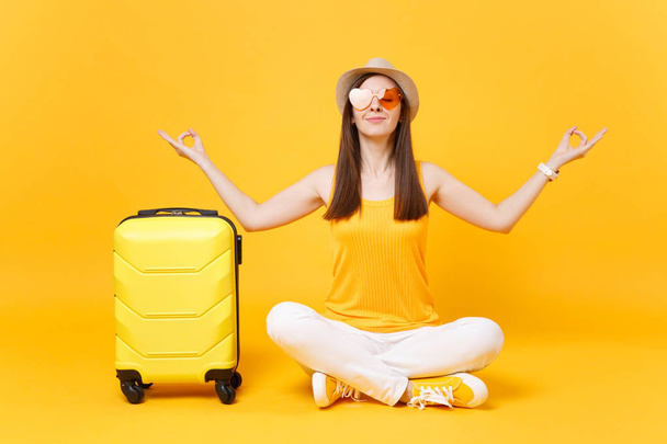 Podróżny turystyczny Kobieta w letni kapelusz siedzieć z walizka skrzyżowane nogi, medytować rozprzestrzeniania się ręce, na białym tle na żółtym tle pomarańczowy. Pasażerów podróżujących za granicę na weekendy wypad. Koncepcja lot Air - Zdjęcie, obraz