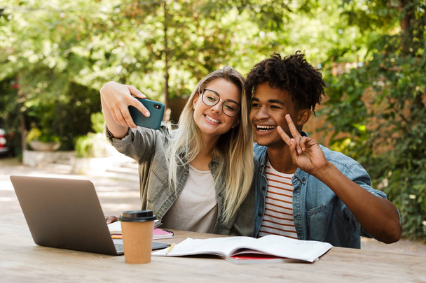 Lovely young multiethnic couple spending time together at the park, studying while sitting at the table with laptop, taking a selfie - Fotografie, Obrázek