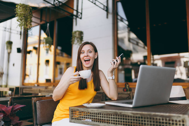glücklich Mädchen im Freien Straßencafé Café sitzt am Tisch mit Laptop PC-Computer, SMS-Nachricht auf dem Handy, trinken Tasse Tee im Restaurant während der Freizeit. Freiberufliches mobiles Bürokonzept - Foto, Bild
