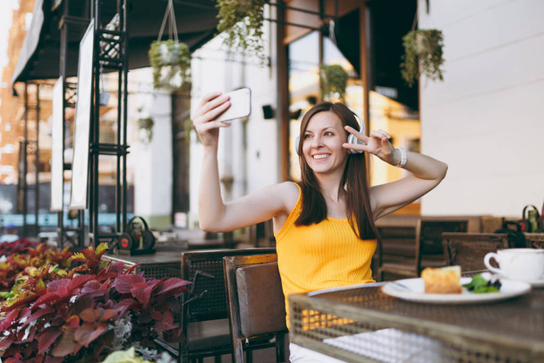 Привлекательная улыбающаяся женщина в кафе на улице сидит за столом, слушает музыку в наушниках, делает селфи на мобильном телефоне, отдыхает в ресторане в свободное время. Концепция отдыха в стиле жизни
 - Фото, изображение