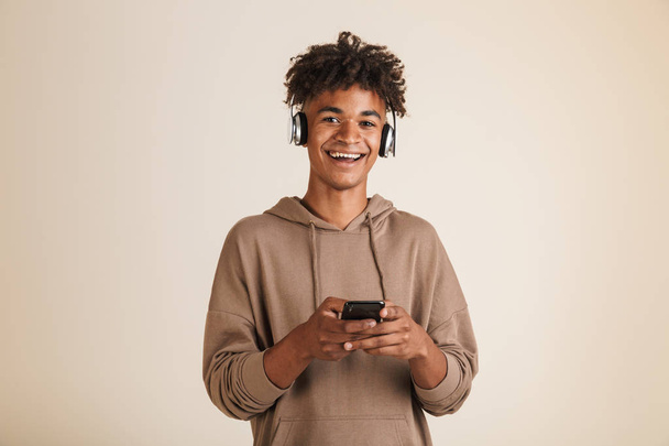 Porträt eines fröhlichen jungen Afroamerikaners in Kapuzenpulli, der isoliert Musik mit Kopfhörern hört und sein Handy in der Hand hält - Foto, Bild