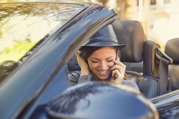 Young stylish woman sitting in modern convertible car and speaking on smartphone happily  - Photo, image