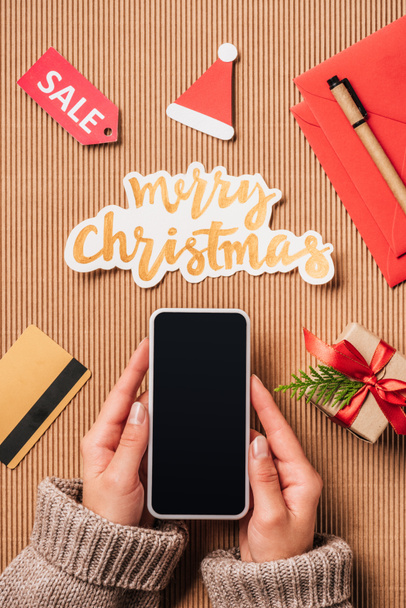 обрізане зображення жінки, що тримає смартфон з порожнім екраном над поверхнею з кредитною карткою, подарунковою коробкою та веселим різдвяним написанням
  - Фото, зображення