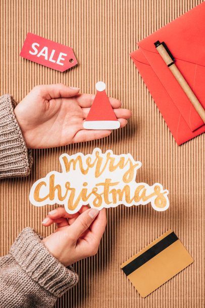 cropped image of woman holding merry christmas lettering and santa hat over surface with credit card and sale  - Photo, Image