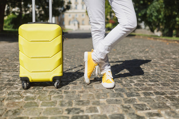 Cropped image traveler tourist woman crossed legs in yellow summer casual clothes with suitcase on road in city outdoor. Girl traveling abroad to travel on weekends getaway. Tourism journey lifestyle - Valokuva, kuva