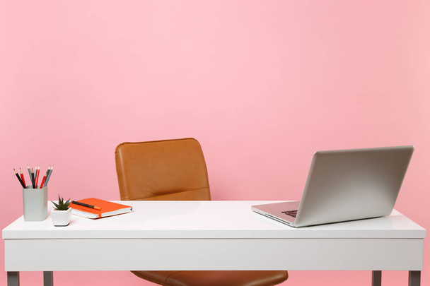 White table, desk with contempopary pc laptop notebook, pencils, Brown leather office chair. Workplace at office isolated on pastel pink background. Office accessories. Copy space for advertisement - Photo, Image