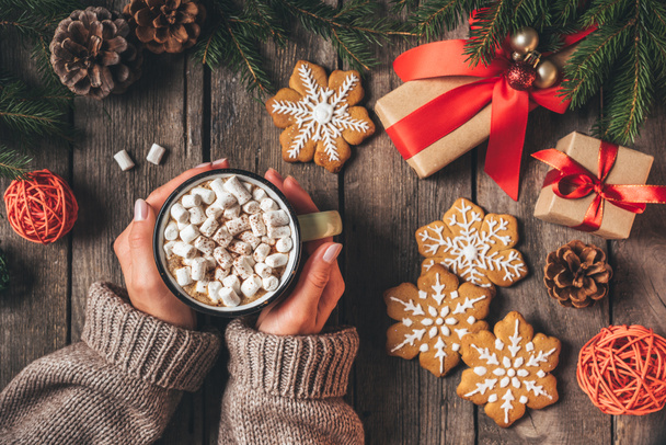 oříznutý pohled ženy držící šálek kakaa s marshmallows a perník na dřevěné pozadí s vánoční dárky - Fotografie, Obrázek