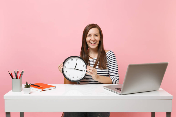 junge, lachende Frau in lässiger Kleidung mit rundem Wecker sitzt an einem weißen Schreibtisch mit zeitgenössischem PC-Laptop auf pastellrosa Hintergrund. Leistung Business-Karriere-Konzept. Kopierraum - Foto, Bild