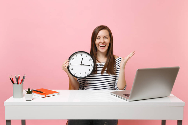 Young cheerful woman holding round alarm clock spreading hands sit work at white desk with contemporary pc laptop isolated on pastel pink background. Achievement business career concept. Copy space - Photo, Image