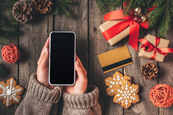 обрізаний вид жінки, що тримає смартфон з порожнім екраном на дерев'яному фоні з кредитною карткою та різдвяними подарунками
 - Фото, зображення