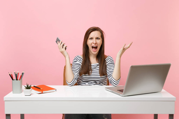 Νέοι ερεθισμένο γυναίκα εξάπλωση τα χέρια ουρλιάζοντας εκμετάλλευση κινητό τηλέφωνο καθμένος εργασία στο γραφείο με φορητό υπολογιστή που απομονώνονται σε παστέλ ροζ φόντο. Επίτευξη της επιχειρηματικής ιδέας καριέρα. Χώρο αντίγραφο - Φωτογραφία, εικόνα