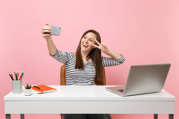 Happy woman doing selfie shot on mobile phone showing victory sign while sit and work at white desk with pc laptop isolated on pink background. Концепция карьерного роста. Копирование пространства
 - Фото, изображение
