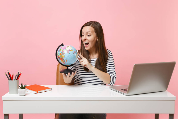 Excited woman looking on world globe planning vacation while sit and work at white desk with contemporary pc laptop isolated on pastel pink background. Achievement business career concept. Copy space - Φωτογραφία, εικόνα