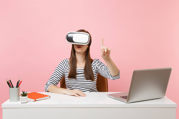 Girl in headset of virtual reality on head touch something like push on button or pointing at floating virtual screen work at desk with laptop isolated on pink background. Achievement business career - Photo, Image