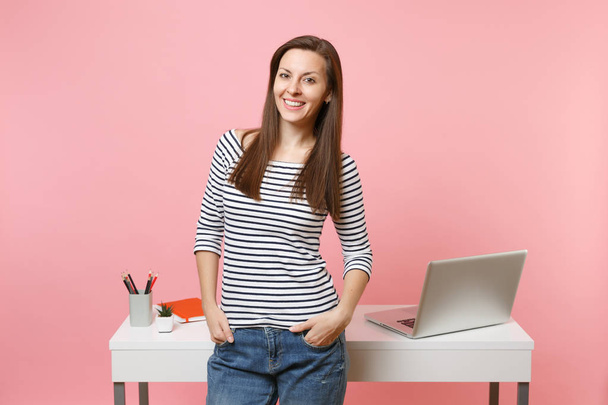 Young successful woman holding hands in pockets work and standing near white desk with pc laptop isolated on pastel pink background. Achievement business career concept. Copy space for advertisement - Photo, Image