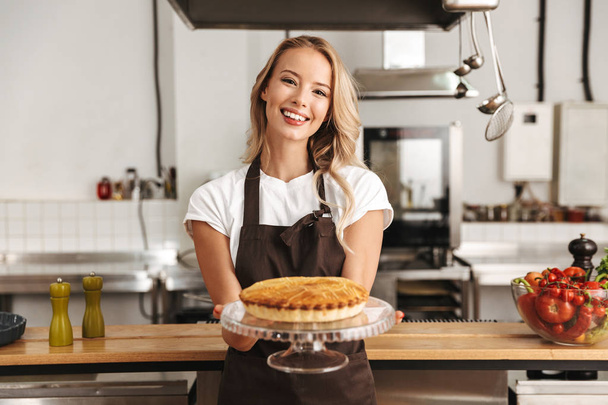 Smiling young woman chef cook in apron standing at the kitchen, showing tasty pie - Foto, afbeelding