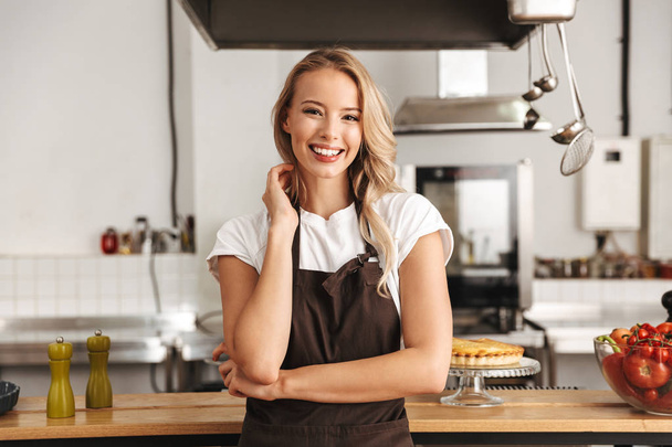 Smiling young woman chef cook in apron standing at the kitchen, looking at camera - 写真・画像