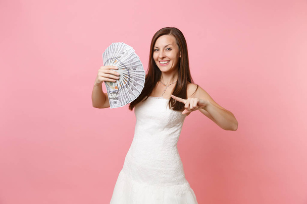 Uśmiechający się panna młoda kobieta w koronki suknia ślubna Biała, wskazując palcem wskazującym na pakiet dużo dolarów, pieniądze na białym tle na pastelowy różowy tło. Organizacja ślubu celebracja koncepcji. Miejsce - Zdjęcie, obraz