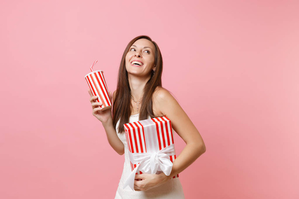 Portrait of cheerful bride woman in wedding dress looking up hold red box with gift, present, plactic cup with cola or soda isolated on pink pastel background. Wedding celebration concept. Copy space - Φωτογραφία, εικόνα