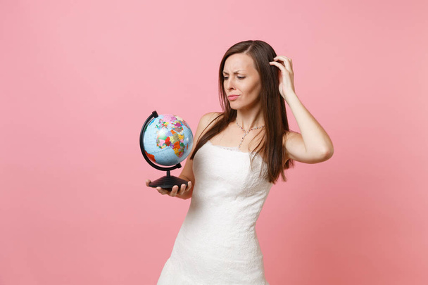Concerned bride woman in wedding dress hold world globe scratching head have problem with choosing place, country for honeymoon, vacation isolated on pink background. Wedding celebration. Copy space - Photo, Image