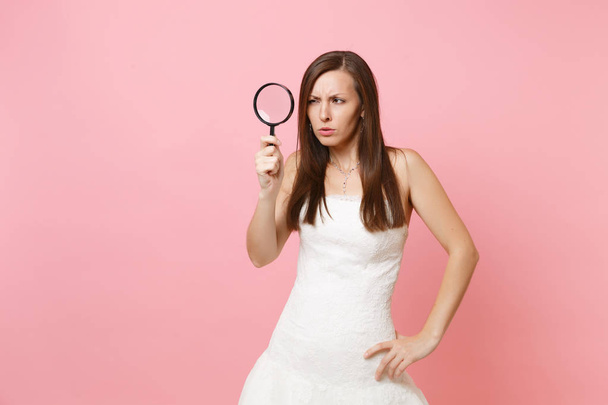Portrait of suspicious bride woman in white wedding dress scrutinizing looking through magnifying glass isolated on pastel pink background. Wedding to do list. Organization of celebration. Copy space - Photo, image