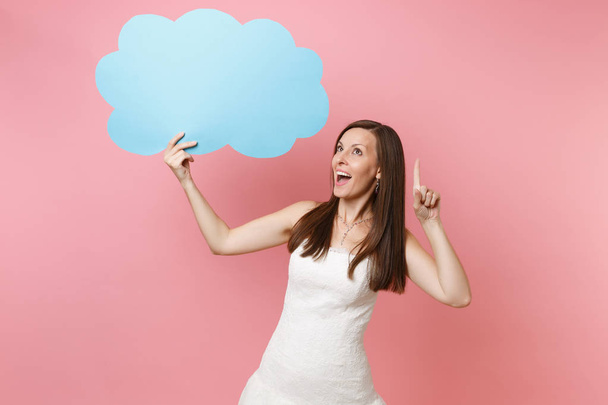 Excited dreamy bride woman in white wedding dress pointing index finger up holding blue empty blank Say cloud speech bubble isolated on pastel pink background. Wedding celebration concept. Copy space - Photo, Image
