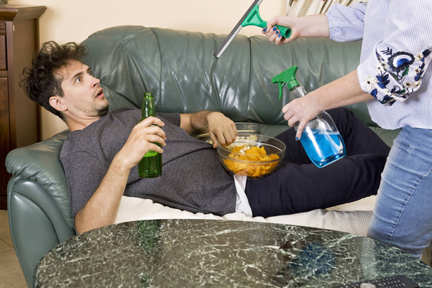 Чоловік п'є пиво і дивиться телевізор, поки його дружина прибирає будинок
 - Фото, зображення