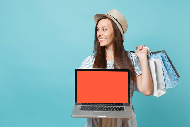 Portrait of woman in summer dress, straw hat holding packages bags with purchases after shopping, laptop pc computer with empty screen isolated on blue pastel background. Copy space for advertisement - Photo, Image