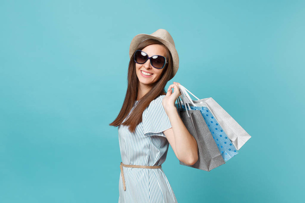 Portrait femme heureuse attrayant à la mode en robe d'été, chapeau de paille, lunettes de soleil tenant paquets sacs avec des achats après les achats isolés sur fond bleu pastel. Espace de copie pour la publicité
 - Photo, image