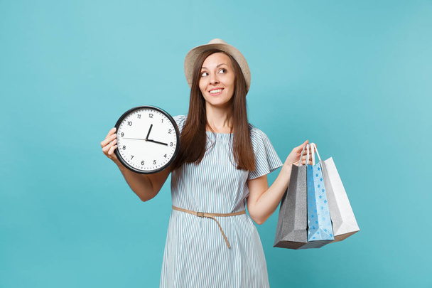 Portrait beautiful young caucasian woman in summer dress, straw hat holding packages bags with purchases after shopping, round clock isolated on blue pastel background. Copy space for advertisement - Фото, изображение