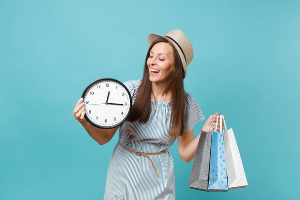 Portrait beautiful young caucasian woman in summer dress, straw hat holding packages bags with purchases after shopping, round clock isolated on blue pastel background. Copy space for advertisement - Fotografie, Obrázek
