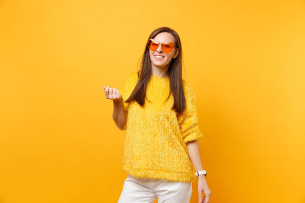 Portrait of cheerful pretty young woman in fur sweater, white pants, heart orange glasses standing isolated on bright yellow background. People sincere emotions, lifestyle concept. Advertising area - Photo, Image