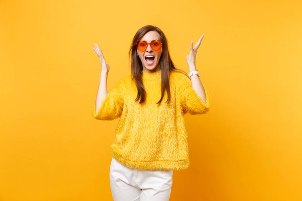 Excited joyful young woman in fur sweater, white pants and heart orange glasses spreading hands isolated on bright yellow background. People sincere emotions, lifestyle concept. Advertising area - 写真・画像